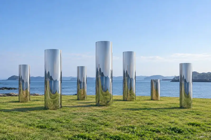 海辺の芝生に、鏡面仕上げのステンレスの円柱が7本立ち、空と海を映している