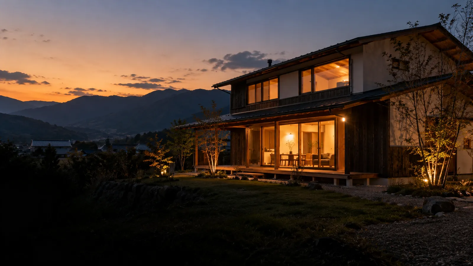 夕暮れ、窓に明かりのともる木造2階建ての家と、背後の山並み